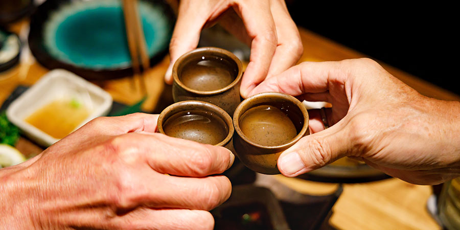 Three people hold small ceramic glasses filled with Sake over their meal. 