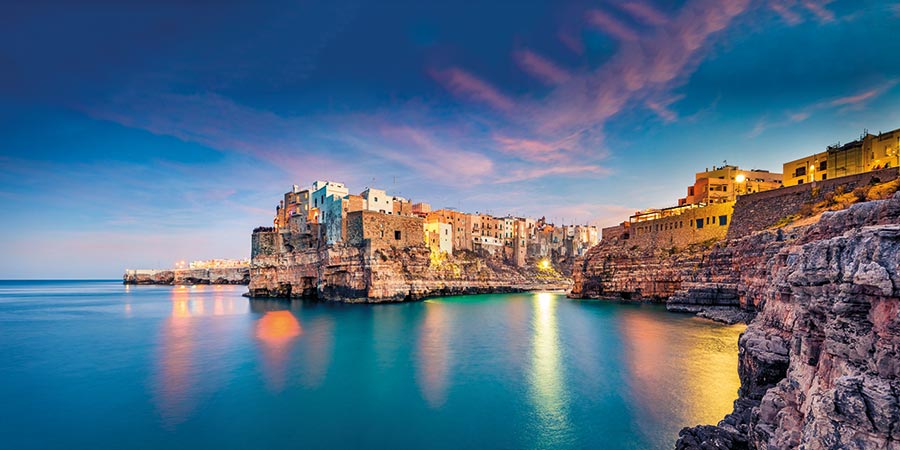 Scenic view of a cove in Puglia's coastline, with crystal-clear blue water meeting a rocky beach, with buildings built into the rocks right up to the shoreline, against a blue and pink sunset sky. 